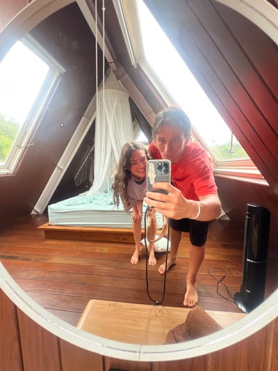 In a round mirror, a man and woman take a selfie. The man wears a red shirt and black shorts, while the woman is in a light purple shirt. They are in a room with wood paneled walls and flooring. Sunlight streams through windows, illuminating a bed with a white canopy. A black heater sits in the background. The phone case held by the man has sticker decorations.
