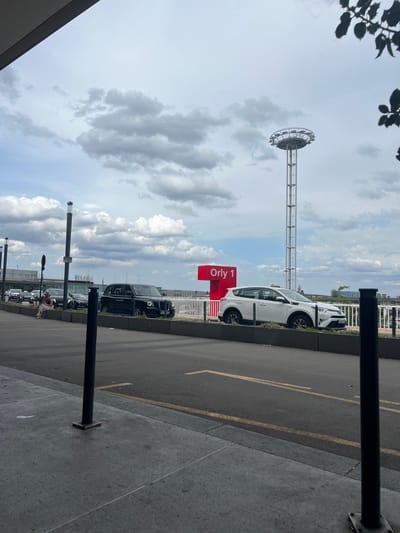This media file captures a scene outdoors, likely at an airport, given the presence of a large terminal building in the background and the sign indicating "Orly 1". The image is oriented vertically, making it appear as if the scene is tilted.

The main subjects include several vehicles, including a white car that appears to be a taxi or ride-share vehicle, and several black cars that resemble taxis. One of these black cars is a distinctive London-style taxi. There are also traffic lines on the asphalt, suggesting a roadway or parking area. In the distance, a tall structure with a circular element at the top, resembling a fairground ride or observation tower, is visible against a cloudy sky.

A prominent red sign with white text reads "Orly 1". The sky is filled with dramatic clouds, suggesting it might be a partly cloudy day. The lighting indicates it is daytime. In the foreground, a person can be seen sitting or resting near the black taxis, and another person is walking further down the sidewalk. The overall setting appears to be a public area outside an airport terminal, possibly a pick-up or drop-off zone. There are black bollards or posts on the sidewalk. The architecture in the background suggests a modern urban or transportation hub environment.