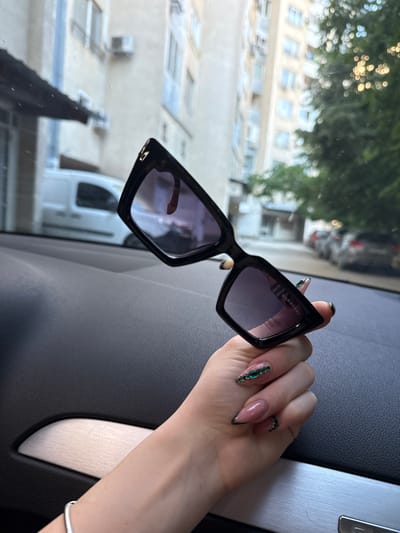 The image shows a woman's hand holding a pair of Burberry sunglasses inside a vehicle. The hand features long, acrylic nails adorned with green jewels and dark tips. The background visible through the car window suggests an urban environment, with buildings and trees. Based on the architecture and foliage visible, the location might be in Plovdiv, Bulgaria, but this is not definitive from the image alone.