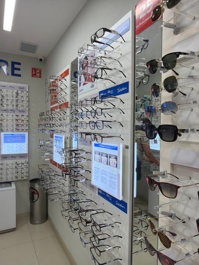 An optical store features sunglasses and prescription eyeglasses displayed on mirrored shelves with pricing and brand signage. Rows of eyeglasses hang from clear plastic stands. Below, framed eyeglasses are arranged on wall-mounted display boards alongside digital informational screens. Price tags include amounts like 1400, 1750, 500, and 888. The brand "Seela" is prominent. A person is reflected in the upper shelf mirror.