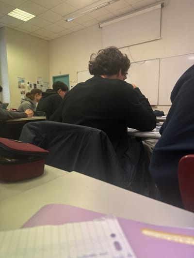 The image depicts a classroom in Albi, France, observed from a low angle, looking towards the front of the room. In the immediate foreground, a light-colored desk surface is visible, bearing a dark red pencil case on the left and a partially visible ruled notebook atop a purple folder on the right.
The central focus is a male individual seated at a desk, seen from behind. He possesses dark, curly hair and wears a black long-sleeved shirt. A dark blue jacket is draped over the back of his chair. He appears to be looking down at his desk. To his left, another male student, also viewed from behind, wears a dark hoodie. Further left and slightly behind the hooded student, a female individual with brown hair, tied back, wears a grey hooded top and is seated at a desk, looking downward. A segment of another individual's arm is visible on the far left. On the far right, the dark blue back and part of a red chair are visible.
The classroom environment includes light-colored walls. The left wall displays multiple papers or posters affixed to it. A light-colored door, possibly teal or light green, is situated on the left wall, near the corner of the front wall. The front wall features two whiteboards and a retractable projection screen positioned above them. A circular clock with a white face is mounted on the front wall between the door and the whiteboards. Overhead, rectangular fluorescent light fixtures are visible on the ceiling. All visible individuals are seated at individual desks.