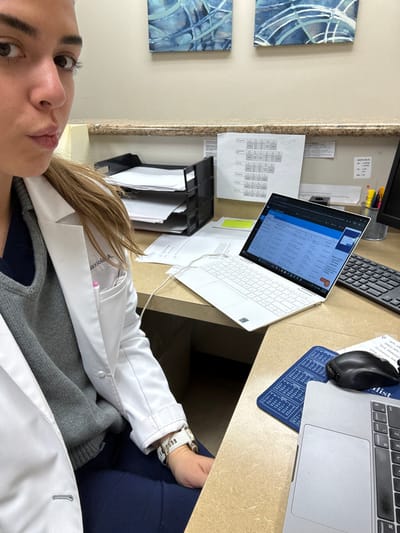 The image is a first-person selfie, taken from a slightly upward angle, capturing a person in what appears to be a professional office or clinical setting. The lighting suggests daytime or a well-lit indoor environment.

The subject of the selfie is a young woman with light brown hair and fair skin. Her face is in the foreground, with her eyes looking directly at the camera. Her expression is neutral. She is wearing a white lab coat over a grey knitted sweater, which is worn over a dark blue or navy top. A pink nametag holder, clipped to the left breast pocket of her lab coat, displays "Dr. Baxiola A." (the surname "Baxiola" is partially obscured, but discernible). A pen with a pink cap is visible in the pocket. On her right wrist, she wears a white wristband, which appears to be a hospital ID or activity tracker, with text reading "ELIGIBLE FOR" and other smaller, unreadable text, along with what looks like a barcode pattern.

The person is seated at a desk or counter with a light brown, possibly laminate, surface. The desk is cluttered with various work-related items. To the left, a black file organizer holds several documents. Behind it, hanging on a light-colored wall or partition, are multiple papers, including a printed schedule or spreadsheet with rows and columns of text and numbers, and a small white note with "NOTICE TO" visible. Another paper has "DCC" and "SPECIALIST" written on it.

On the desk, there are several electronic devices:
- A white Dell XPS laptop is open and positioned centrally, displaying what appears to be a web browser or application with a blue header and content showing lists or tabulated data. A small 'P' logo is visible in one of the tabs. A white charging cable extends from the laptop.
- A black computer keyboard is partially visible behind the white laptop.
- To the right, a grey MacBook laptop is also open, with its keyboard and trackpad visible.
- A black Logitech computer mouse rests on a blue mousepad, which features a white grid pattern resembling a Braille reference chart. The mouse is positioned near the MacBook.

On the wall to the left of the desk, two square pieces of abstract art are displayed. They feature blue and white patterns, possibly watercolor or acrylic designs, adding a decorative touch to the workspace.

The overall impression is that of a busy professional environment, possibly a doctor's office or administrative area, where the individual is taking a brief moment to capture a selfie amidst her work.