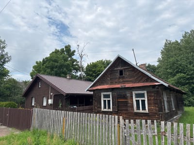 The image captures a view of two houses in Trześcianka, Poland, under a cloudy sky. The foreground features a weathered wooden fence with vertical planks. To the right, a rustic, dark brown wooden house with a red metal roof is prominently displayed. This house has several windows, some with white frames and shutters, and shows signs of age. To the left, a more modern, dark brown house with a tiled brown roof is visible. This house has a few windows and a visible house number "96" along with an outdoor unit for air conditioning. Both houses are surrounded by lush green trees and vegetation. The overall scene depicts a rural or semi-rural setting with a blend of older and newer structures.