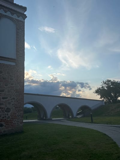 The image captures a view of the Zhykhavichy Castle bridge, an architectural landmark in the Zhukhavichy rural council of Belarus. The bridge, with its three distinct arches, spans across a green landscape. On the left, a portion of a red brick building with stone accents and decorative white trim is visible, suggesting historical architecture. The sky above is a blend of blue with scattered clouds, some of which are illuminated by sunlight, creating a dramatic effect. A lone tree stands to the right of the bridge, its branches reaching towards the sky. A paved walkway curves through the grassy area in front of the bridge, with lampposts lining the path. There are no people clearly visible in the scene.