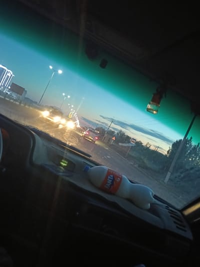 The image is a photograph taken from inside a vehicle, looking out through the windshield at dusk. The sky is a gradient of deep blue at the top to lighter hues near the horizon, with a few wispy clouds. The scene depicts a road with traffic, illuminated by streetlights and headlights.
In the foreground, the dashboard of the car is visible, with a white bottle of Fanta resting on it. A fuzzy object, possibly a car air freshener, hangs from the rearview mirror.
Outside the car, several vehicles are on the road. The headlights of approaching cars are bright, creating streaks of light due to motion or camera settings. On the left side of the road, there's a modern building with bright, vertical lights. Streetlights are lining the road, casting a warm glow.
In the middle ground, there are trees and signs along the roadside. One sign appears to be a yellow triangle with a black symbol, indicating a hazard or warning. Another sign is blue with white markings, possibly a bus stop sign.
The overall impression is of a drive during twilight on a road that seems to be in or near a populated area, potentially a city or town. Given the context provided, this scene is located in Kungrad, Uzbekistan. The road appears to be a multi-lane highway or a major street with street lighting and surrounding infrastructure. The presence of traffic suggests it's an active time of day.