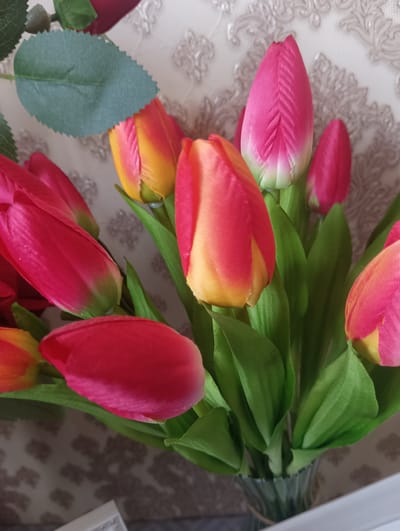 This is a close-up, eye-level, indoor photograph of a bouquet of artificial tulips in a glass vase. The tulips are vibrant, with some being a bright pink and others a gradient of red and yellow. Green artificial leaves are interspersed among the flowers. The background is a textured wallpaper with a damask pattern in muted tones. The lighting is soft, creating subtle shadows. There are no people or animals visible in the image. The scene appears to be a still life arrangement. The provided information does not include any details about the city of Tallinn, Armenia, as the photograph is not related to that location.