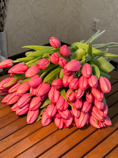 A large bouquet of approximately 50-60 vibrant pink, tightly closed tulip buds with prominent green stems and leaves is centrally displayed on a wooden slatted table. The table exhibits a medium-brown hue and a horizontal grain pattern. The tulips are arranged in a dense cluster, filling the majority of the frame, with their stems extending upwards and to the right. In the background, a light-colored wall with a subtle, repeating patterned wallpaper is visible. A white, double electrical outlet is present on the wall in the upper right quadrant. A portion of a tall, slender object, possibly a decorative vase with a dark, vertical striped or textured pattern, is visible in the upper left corner. The lighting appears to be ambient, highlighting the texture of the tulips and the wooden surface. No people are present, and no specific actions or interactions are depicted. The interior environment provides no discernible visual cues to identify a specific location within Slavyansk-na-Kubani, Russia.