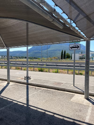 The image depicts an outdoor scene under a shaded structure, likely a parking area or rest stop. A dark grey fabric shade, supported by metal poles, covers the foreground. Beyond the shade, a paved road is visible, separated by a metal guardrail from a grassy embankment. In the background, a mountain range dominates the horizon, with trees and some buildings dotting the landscape. A road sign with a black car silhouette on a white background is posted on a pole to the right. The presence of the guardrail and road suggests a location near a highway or major road. The overall environment appears to be a sunny day. Based on the visual context, the location can be identified as being in the Municipal Unit of Iera Poli Messologhiou, Greece, likely near a transportation hub.