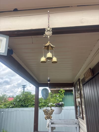 The image captures an outdoor scene under a pale, wood-paneled overhang, likely a porch or veranda. A wind chime, featuring an owl at the top, three brass bells, a black bead, and a butterfly charm at the bottom, hangs from the ceiling. To the left, a modern LED floodlight is attached to the overhang. Beyond the overhang, a garden with lush green trees, a red-roofed structure, and a corrugated metal fence is visible under a cloudy sky. A string of globe lights is strung across the garden area. On the right side of the image, a wooden door with vertical planks is partially visible, with a carved wooden sign that reads "BATHS." A white planter with purple flowers hangs in the background. The overall setting suggests a residential outdoor space in Лотос Снт (марьино), Russia.