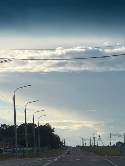 The image depicts a road scene in the vicinity of Бальшавіцкі сельскі Савет, Belarus. The viewpoint is from a vehicle traveling on a paved road. In the foreground, the road has visible lane markings. Along the left side of the road, a row of tall, dark lampposts with curved arms are spaced at intervals. Beyond the lampposts, there is a line of dense, dark green trees. Some low-lying buildings are partially visible amongst the trees on the left.
The road leads into the distance, curving slightly uphill. Further down the road, a light-colored car is visible. On the right side of the road, there are more lampposts, some with electrical wiring or utility poles. A bus stop sign is also present on the right.
The sky above is a gradient of blue, transitioning from a darker teal at the top to a lighter sky blue towards the horizon. Scattered across the sky are numerous clouds, some appearing as thin, wispy streaks and others as more substantial, puffy formations. The overall lighting suggests it is daytime, possibly late afternoon or early evening, given the soft light and cloud cover. Power lines are strung across the upper portion of the image, running parallel to the road.