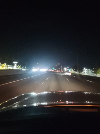 The image displays a nighttime perspective from inside a vehicle traveling on a multi-lane highway or freeway in Fife, United States. The top portion of the vehicle's dashboard or hood is visible at the bottom of the frame, showing reflections of interior lights on the windshield. The roadway ahead features multiple lanes delineated by white dashed lines, with a solid white line marking a shoulder to the left.

Moderate vehicular traffic is present. Numerous distant headlights illuminate the lanes ahead, with a distinct white sedan visible in the right-hand lane. Headlights and taillights from oncoming vehicles are visible in the opposing lanes, separated by a median or barrier.

The environment is artificially lit. Along the left side of the highway, illuminated commercial buildings and signage are discernible. On the right, streetlights are active, and a distant green highway sign with white text is visible above the roadway. The sky overhead is dark, indicating a nocturnal setting. No individuals are directly visible.