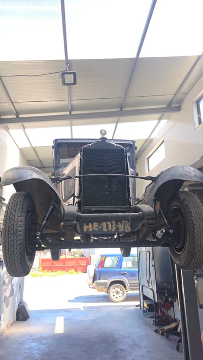 The image depicts the underside of a vintage vehicle, possibly a truck, elevated on a mechanical lift inside a garage. The vehicle features a radiator, front tires with a classic tread pattern, and a suspension system with visible leaf springs and shock absorbers. Yellow lettering, "HE-111-NB," is painted on the engine component. In the background, a blue van is visible, parked inside the garage. Sunlight streams through overhead windows. The location is identified as Balgarevo, Bulgaria.