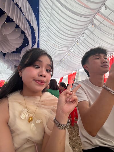 This image captures two young individuals posing at an indoor or covered outdoor event, likely in Tay Son Commune, Vietnam.
In the foreground, a young woman with long dark hair is looking directly at the camera, wearing a light-colored, possibly cream or light yellow, draped garment. She has a subtle smile or pout and holds her left hand up, forming a peace sign. Her left wrist displays a silver-colored watch with a link bracelet, and around her neck, she wears a delicate gold chain necklace with two gold rings as a pendant, resting on her chest.
Behind her and slightly above, a young man with short dark hair is visible, wearing a plain white t-shirt. He has a silver-colored link bracelet on his left wrist and is looking upwards and to his left with a neutral expression.
The setting appears to be a decorated venue, possibly a large tent or event hall. The ceiling is draped with white fabric, creating a soft, pleated effect, and small lights are interspersed within the drapes. In the background, there are sections of blue fabric decorated with white ruffled patterns and more small lights, as well as red and white fabric decorations further back. Other indistinct figures are visible in the distance, suggesting a gathering or celebration. The lighting indicates it is daytime, with bright ambient light illuminating the space. No visible text is present.