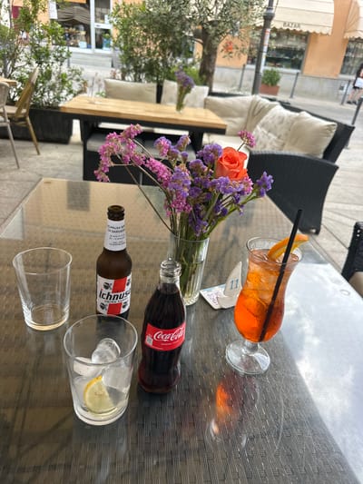 An outdoor table at a cafe in Villasimius, Italy, is set with drinks and a floral arrangement. The table surface is textured and reflective, with the dappled light of an overcast day. On the table, a bottle of Ichnusa Birra di Sardegna beer, a bottle of Coca-Cola, and a glass filled with ice and a lemon slice are visible. A vibrantly colored orange cocktail with a straw and an orange slice garnish is also present. A vase of purple and pink dried flowers, with a single orange rose, adds a decorative element. In the background, outdoor seating with cushions and potted plants are seen, along with a storefront with awnings and the word "BAZAR" visible. A woman in dark clothing and a white top is walking in the distance.