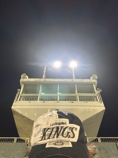 A low-angle, upward shot captures a tall, concrete structure featuring two stories, each with barred windows. Bright stadium lights shine above, casting a glare. A baseball cap with "Los Angeles KINGS" prominently displayed obscures part of the view, suggesting the photographer's perspective. The night sky is dark, providing contrast to the brightly lit structure and hat. A section of metal fencing is on the far right edge.