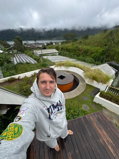 A man in a gray hoodie and sweatpants stands barefoot on a wooden deck, seemingly on a balcony overlooking a lush, green landscape. The backdrop features a mist-shrouded lake and mountains. Manicured gardens and unique architectural elements surround him. The hoodie is adorned with various patches and designs. The shot is taken from a high angle, emphasizing the steep drop of the landscape.