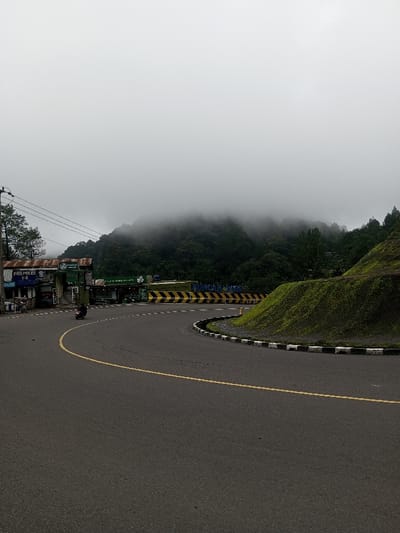 This image displays a curving asphalt road in the city of Deothang, Bhutan, under an overcast sky with mountainous terrain partially obscured by fog. The road, featuring a yellow centerline, transitions into a sharp left turn. Along the outer edge of this curve, a black and white striped curb borders a steep, verdant embankment. To the left of the road, a series of structures with corrugated metal roofs are visible, including a building prominently marked "POS POLISI T-B" in white text on a blue sign. Adjacent to these buildings, a set of yellow and black striped barriers extends across the area, indicating a checkpoint. A utility pole with multiple power lines is situated on the far left. A single person on a dark scooter/motorcycle is depicted on the road, mid-turn, moving leftward. The background reveals dense green forests ascending into mountains largely enveloped by thick mist.