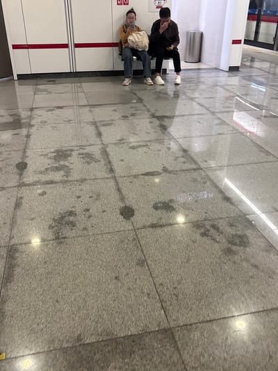 Two people are sitting against a white wall in what appears to be a subway station or similar public space. One person, wearing a brown jacket and black pants, is bent over. The other person, wearing a yellow jacket and blue jeans, is looking at a phone. There's a silver trash can nearby. The floor is tiled and has a wet or stained appearance. The space is illuminated by overhead lights.