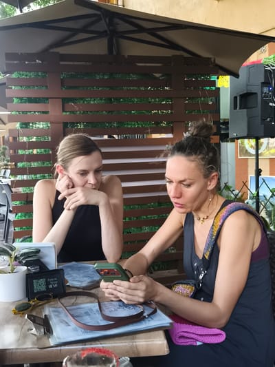 Two women are seated at a wooden table at an outdoor cafe. The woman on the left is fair-skinned, with blonde hair pulled back, wearing a black sleeveless top. She rests her head on her hands and looks thoughtfully at the table. The woman on the right has her hair in a bun and is wearing a dark sleeveless dress with a beaded shoulder strap. She is holding and looking at a cell phone with a green case. On the table are a potted plant, a check presenter with a QR code, yellow sunglasses, a clipboard with papers, a brown strap, and other indiscernible objects. The backdrop includes a wooden lattice fence with green foliage visible behind it. A speaker and artwork are visible on the right side of the frame. This image likely was taken in the city of Querétaro, Mexico, as the QR code in the photo is on an item referencing "Fonda del Refugio Menu" which is in Querétaro, Mexico.