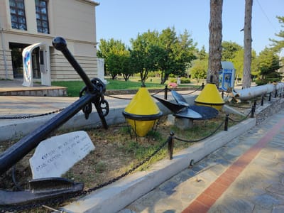This image depicts an outdoor exhibit of naval artifacts in Litochoro, Greece. In the foreground, a large, black anchor rests on the ground, with a white stone slab placed in front of it. The stone has text inscribed on it. To the right of the anchor, two bright yellow buoys are displayed, each with a black band around its middle and a black top. Behind the buoys, a propeller and a torpedo are visible, suggesting a maritime or naval theme for the display. The objects are separated by black metal chains and posts.
The setting appears to be a park or an outdoor museum area adjacent to a building with light-colored walls and dark windows. Trees and green grass are visible in the background, indicating a pleasant, possibly sunny day. The sky is clear and blue, suggesting it is daytime. The overall atmosphere is one of historical display and remembrance, with the artifacts presented as points of interest.
Visible text on the white stone slab appears to be in Greek. The larger inscription reads "ΕΧΟΥΜΕ ΤΗΝ ΚΑΙ ΠΑΤΡΙΔΑ ΟΤΑΝ ΕΧΟΥΜΕ ΠΛΟΙΑ ΣΤΗ ΘΑΛΑΣΣΑ", which translates to "WE HAVE THE HOMELAND WHEN WE HAVE SHIPS AT SEA". Below this, smaller text is present, possibly names or dates. A blue and white telephone booth is visible further in the background, near some trees.
