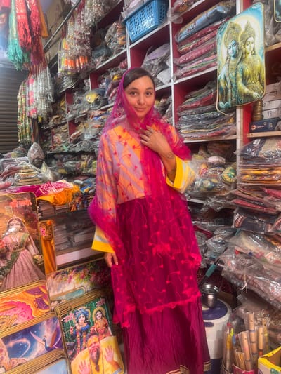 A woman wearing a traditional Indian dress and head covering stands in a shop filled with religious merchandise in Mathura, India. The shop has shelves stocked with packaged fabrics and other items. The woman is fair-skinned, and the translucent pink head covering partly obscures her face. Her dress is a combination of yellow, lavender, and dark pink. Her left arm is extended, showing tattoos. The foreground is dominated by a stack of framed religious images. A framed depiction of Hindu deities is visible on the upper left.