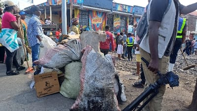 This image depicts an outdoor urban scene in Gisenyi, Rwanda, centered on what appears to be a community cleanup operation or public gathering. In the immediate foreground, a significant pile of collected debris is visible, contained within various large sacks of different colors (grey, white, reddish-brown, green). A cardboard box, partially open and labeled "SERENGETI LAGER," rests on the ground near these sacks. To the far right foreground, the lower body and right arm of an individual in tan pants are visible, holding a black camera mounted on a tripod or monopod. The ground consists of rough, uneven dirt and scattered debris.

In the midground, numerous individuals are present. On the left, a person wearing a pink shirt, dark pants, and black sandals holds an orange-handled broom. Another person to their right, wearing a blue and white checkered shirt and white gloves, stands near more sacks. Further into the midground, a substantial concrete or stone block is positioned amongst the debris. A diverse group of people, varying in age and attire, are scattered across the scene. One individual is discernible wearing a bright yellow reflective vest over a dark shirt, holding a stick-like object.

The background features a row of buildings with distinctive, vibrant murals displaying geometric designs and the brand name "PRIMUS" in prominent lettering on at least two sections. The sky is bright, suggesting daytime conditions. The overall scene indicates organized public activity, possibly a civic cleanup or community event, with elements of media documentation.