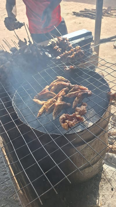 The image captures an outdoor scene in Kporva, Nigeria, where meat is being grilled over an open fire. The primary subjects are skewers of meat, some appearing to be chicken and others resembling small whole fish, arranged on a metal grill. A person wearing a red shirt is partially visible on the left side of the frame, seemingly tending to the grill. Wisps of smoke rise from the cooking meat, indicating the grilling process is in full swing.

The setting appears rustic, with a dirt or sandy ground and what looks like a large clay pot or drum serving as the base for the grill. The overall atmosphere is one of casual outdoor cooking, possibly for a meal or a market stall. The lighting suggests it is daytime, with natural sunlight casting shadows. There are no visible text elements in the image. The focus is on the food preparation and the traditional grilling method being employed. The scene conveys a sense of activity and the preparation of food in a communal or informal setting.