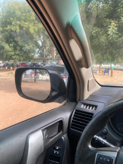This image depicts a scene observed from the interior of a stationary vehicle in Yan Dala, Nigeria, looking forward and left.

**Interior:** The immediate foreground and right side display components of the vehicle's interior. A dark, worn steering wheel is partially visible on the far right. The dashboard, dark gray with a horizontal air vent, is situated beneath a light beige A-pillar featuring an integrated grab handle. The door panel, also dark, includes a silver-colored door handle and a cluster of black power window and door lock switches.

**Exterior (Forward View):** The view outside the vehicle reveals an open area with reddish-brown earth ground. Numerous green-leafed trees are scattered across the landscape under a clear, bright sky. In the mid-ground, several vehicles are parked, including a red sedan and multiple dark-colored vehicles. Various colorful structures, possibly benches or decorative elements, painted in blue, green, and white, are visible in the background, suggesting a park or public recreational space. No individuals are distinctly visible in this forward view.

**Exterior (Reflected in Mirror):** The driver's side exterior mirror, prominent on the left, reflects the scene directly behind the vehicle. This reflection shows more of the reddish-brown ground and a light-colored building with distinct vertical striped decorations (blue, red, orange, white) on its facade. Additional green trees and a partial view of another light-colored vehicle are also visible in the mirror's reflection. No individuals are distinctly visible in the reflection.