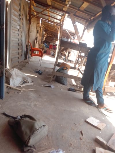 The image displays an indoor or covered outdoor market in an unspecified city within Nigeria, identified as 0801712591. The setting features a rough concrete floor covered with scattered sawdust, wood fragments, and plastic debris. Overhead, a makeshift roof structure consists of wooden beams and supports, topped with corrugated metal sheets, allowing natural light to penetrate.
In the foreground, an olive-green canvas shoulder bag rests on the floor to the left. In the mid-ground left, an empty orange plastic chair with silver legs is positioned against a wall constructed from vertical wooden slats or shutters. Near the chair, a large, crumpled light-colored sack lies on the floor, alongside a pair of dark flip-flops.
A person, likely male, is standing in the right mid-ground, facing away from the camera and slightly to the left. They wear a light blue tunic over dark trousers and light-colored flip-flops. Their right hand is raised towards their head, obscuring part of their face. Adjacent to this individual, a wooden market stall structure is visible, with a white bowl placed on its upper surface. Below the stall, a dark-colored sack and a yellow plastic jerrycan are present.
In the background, a long market aisle extends into the distance, lined with numerous wooden stalls. Multiple individuals are seated or crouched near these stalls, appearing to engage in transactions or market activities. Red plastic crates are discernible among the merchandise displayed on the stalls. The overall scene suggests a functional, informal market environment.