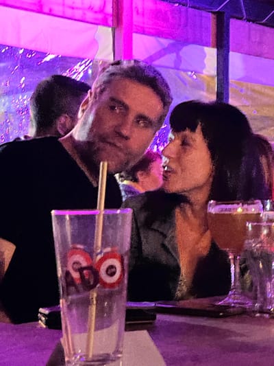 The image captures an indoor scene, likely at an evening event, dominated by vibrant artificial lighting. The photo appears to be rotated 90 degrees clockwise from its natural upright orientation.
Two main individuals are visible. In the foreground, positioned on what would be the left side if the image were upright, is a man with light skin, short dark hair, and a short beard. He is wearing a dark top and looks directly towards the viewer with a neutral expression. He is holding a transparent rectangular holder containing two wooden skewers. Each skewer holds a circular, target-like object featuring concentric rings of red, white, and black. One of these objects also displays an ambiguous dark marking that could be a stylized number "8" or letter "G". The top object also seems to have a similar ambiguous marking. A third person's head is partially visible behind the man's left shoulder.
In the mid-ground, on what would be the right side of the upright frame, a woman is seen. She has dark hair styled in a bob cut, light skin, and wears a dark top. Her head is tilted slightly downwards, and she appears to be looking towards the right, seemingly engaged with something outside the frame.
The background features bright, colorful illumination, primarily in shades of purple and pink, with hints of yellow-orange. These lights illuminate what appears to be a wall or decorative panels, contributing to a lively, possibly festive, atmosphere. No explicit text is clearly legible beyond the ambiguous markings on the skewered objects. The scene suggests a social gathering or entertainment venue in Saint-Priest-en-Jarez, France.
