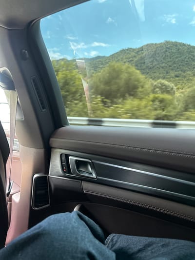 An interior shot from a car reveals an open sunroof framing a scenic view of a green mountain range under a bright, partly cloudy sky. The car's interior features a luxurious design with brown leather trim, metallic accents, and visible door handle details. The image captures a sense of motion, suggesting the car is driving through this picturesque landscape. A person is visible to the right.