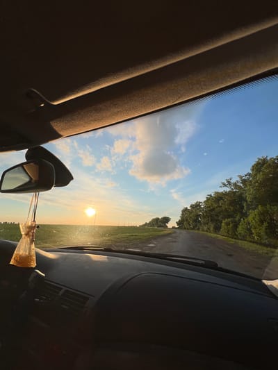 The image captures a first-person perspective from within a vehicle, looking forward through the front windshield towards a rural landscape at sunset in Tovstoluh, Ukraine. The sky, a clear blue with scattered white cumulus clouds, features a visible crescent moon in the upper right quadrant. The sun, a bright, intense orb, is low on the horizon, positioned slightly left of center, casting warm light and reflections on the windshield and dashboard.

The vehicle's dark headliner is visible at the top of the frame, alongside the top edge of the windshield which includes a dotted frit band. A black rearview mirror is centrally positioned, with a small, translucent pouch containing granular, yellowish material suspended from it, likely an air freshener or charm. The dark dashboard occupies the lower portion of the frame, showing air vents and contours, with parts illuminated by the setting sun.

Outside the vehicle, an unpaved dirt road, appearing wet or muddy in patches, extends into the distance, bordered by vegetation. To the left of the road is a vast green field, possibly agricultural, with indistinct structures or poles visible in the far distance. To the right, a dense line of trees with lush green foliage parallels the road. No individuals are visible in the scene.