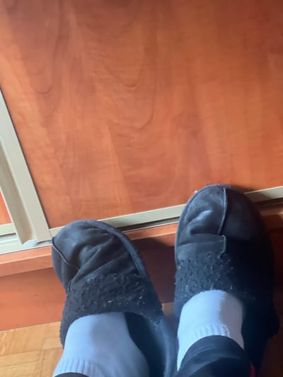 The image displays a downward view of a person's lower legs and feet, situated indoors in Ittenheim, France. The subject's feet are clad in white socks and black, textured slippers, which exhibit visible pilling on their outer surfaces. The immediate ground surface is composed of reddish-brown wood with a discernible grain. A light-colored, horizontal trim or frame element extends across the scene, separating the floor area from a vertical wooden panel in the background. No other individuals, actions, or interactions are observable.