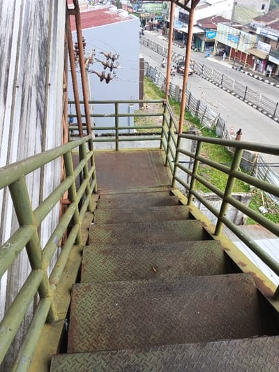 An elevated, downward-angled view captures a section of a metal staircase and an urban streetscape in Kidapawan, Philippines. In the foreground, a flight of industrial-style stairs, constructed from diamond plate metal, exhibits significant weathering, rust, and a faded olive-green and brown coloration. The stairs are flanked by green metal railings, also weathered. A single cigarette butt lies on the third visible step from the bottom.
To the left, a corrugated metal wall, likely part of an adjacent building, is partially visible. At the top of the stairs, a flat metal landing is present, bordered by further green metal railings. Beyond this landing, an array of ceramic insulators on a utility structure supports overhead electrical lines.
The midground and background depict a city street. A two-lane asphalt road with a concrete median barrier spans horizontally across the frame. Multiple vehicles, including motorcycles and at least one orange-brown car, are present on the road. Pedestrians are visible on the sidewalks adjacent to the road. A row of various low-rise commercial buildings, featuring different architectural styles, colors (including light blue and white), and roof types (red corrugated), lines the street. Patches of green grass and sections of wire fencing are situated between the foreground structure and the main road. Distant commercial signs and billboards are also visible under an ambient daylight sky.