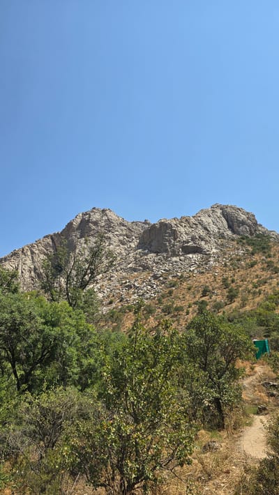 This image depicts a mountainous landscape with a clear blue sky. In the foreground, there are various types of vegetation, including trees and shrubs. A dirt path is visible, winding its way up the side of the hill. Further in the background, a large, rocky mountain rises, its slopes covered in scree and sparse vegetation. A small, bright green structure, possibly a shelter or building, is situated on the hillside. The environment appears to be dry and arid, with a sense of remoteness. The scene is devoid of any people or animals. The image suggests a natural, outdoor setting in a region with significant elevation changes.