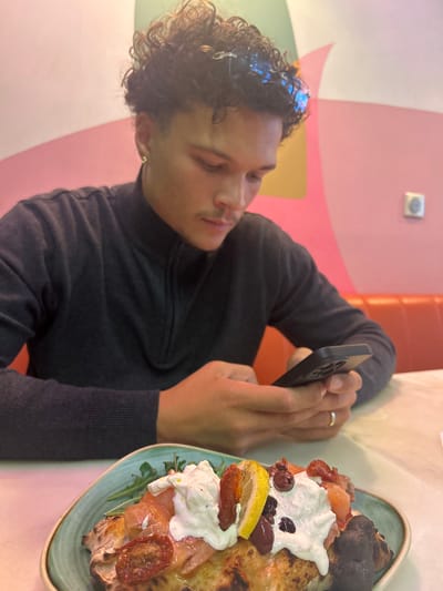 A person with dark, curly hair and a mustache is sitting at a table, holding a black smartphone and appearing to take a photograph of a plate of food. The food, placed on a blue-green plate, includes what seems to be pizza topped with white dollops, tomatoes, olives, a lemon slice, and burnt crust. The table has a light-colored surface, possibly marble or a similar material. The background includes a wall with colorful, curved patterns in shades of pink, orange, and yellow. There is a glimpse of an orange cushioned seating area in the background, and a light switch is visible on the wall. Based on the description in the image title, the location is likely in Serris, France.