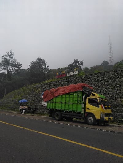 An overcast and foggy roadside scene in Cugenang, Cianjur Regency, Indonesia, is depicted. The location is identified by a large, colorful sign on a steep hillside reading "Cianjur Bangkit." A two-lane asphalt road with a double solid yellow center line occupies the foreground. On the right side of the road, a large retaining wall constructed from dark stones supports a vegetated embankment.
A yellow and bright green cargo truck, heavily loaded with goods covered by a red tarp, is parked on the shoulder parallel to the retaining wall. Further down the road from the truck, a small vendor cart with a blue umbrella is positioned against the retaining wall. One individual is seated behind the vendor cart, facing away from the camera. A second individual is partially visible between the vendor cart and the truck, near the base of the retaining wall. Dense trees and foliage cover the hillside, and a communication tower is partially visible through the fog on the distant right horizon.