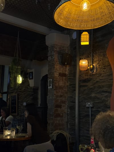This image shows a dimly lit interior, likely a restaurant or bar, in Windermere, United Kingdom. The walls appear to be constructed of stone or brick. Various light fixtures are visible, including a large woven lampshade, a small decorative sconce with a checkered pattern, and another light fixture with a ribbed globe. A metal shelf runs along the wall, supporting various items. The image also captures portions of people: one person is seated at a table with drinks, and another individual is visible in the lower portion of the image. A small chalkboard with handwritten text advertising "Soup of the day" and a "Cocktail of the moment" is present. The overall atmosphere appears to be dark and intimate.