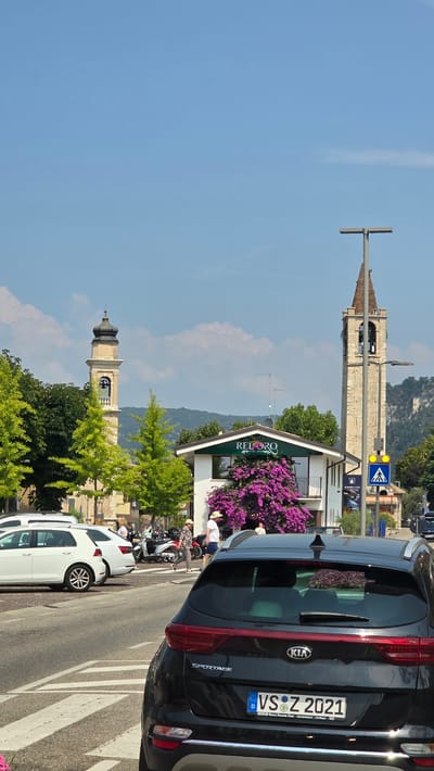 The image depicts a street scene in Bardolino, Italy. In the foreground, a black Kia Sportage is parked facing the viewer. The car's license plate reads "VS Z 2021". Behind the Kia, a white Volkswagen Golf and another white car are parked. Further back, several motorcycles are visible. Two white buildings stand side-by-side; the one on the left has a clock tower and the one on the right is adorned with vibrant purple bougainvillea. A tall, stone bell tower, characteristic of Italian architecture, rises on the right side of the image, with a cross at its peak. Several people are seen walking and standing on the sidewalk in front of the buildings. Lush green trees line the street, and rolling hills are visible in the distance under a clear blue sky with scattered clouds. A street lamp and a pedestrian crossing sign are also present.