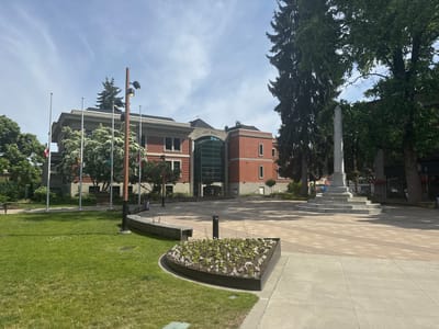 This media file captures a bright, daytime scene of a civic square or plaza in front of a City Hall building in Port Coquitlam, Canada.

In the foreground on the left, a well-maintained grassy area is visible, partially bordered by a modern-style wooden bench. Adjacent to the grass is a dark, curved planter box filled with small white and light purple flowers. The ground then transitions to a large, patterned paved plaza, leading towards the main building. A tall, dark brown pole with three spotlights stands in the mid-left of the plaza.

The central subject is a multi-story building identified by the text "CITY HALL" above its prominent central entrance. The building features red brick construction, with the main entrance being a tall, modern glass-enclosed atrium. Several windows are visible on the building's facade. To the left of the City Hall, a row of flagpoles stands, with some flags, including what appears to be a Canadian flag, flown at half-mast.

To the right of the paved plaza, a tall, light-colored stone monument with an obelisk-like shape stands on a tiered base. Large, mature trees with dense green foliage frame the scene on both sides, including tall evergreen trees on the right. The sky overhead is bright blue with scattered, wispy white clouds, indicating sunny weather. In the far background, beyond the monument and trees, a faint sign with the word "SOUP" is visible on a distant storefront, hinting at commercial establishments nearby. The scene depicts a peaceful and well-maintained public space.
