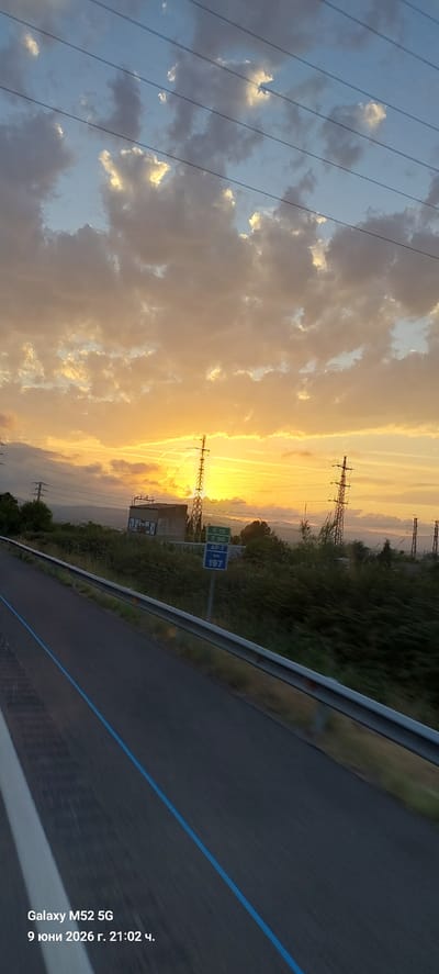 This is a landscape photograph taken on a highway, possibly the AP-7, in the municipality of Olèrdola, Spain, at sunset. The sky is filled with dramatic clouds illuminated by the setting sun, casting a warm orange glow. Power lines and electricity pylons are visible across the scene. In the foreground, the asphalt road with its white and blue markings curves to the left. A metal guardrail runs along the side of the road. A blue road sign indicates "AP-7" and "197 km". In the middle ground, there are some buildings and dense vegetation. The timestamp "9 юни 2026 г. 21:02 ч." and "Galaxy M52 5G" are watermarked at the bottom left corner.