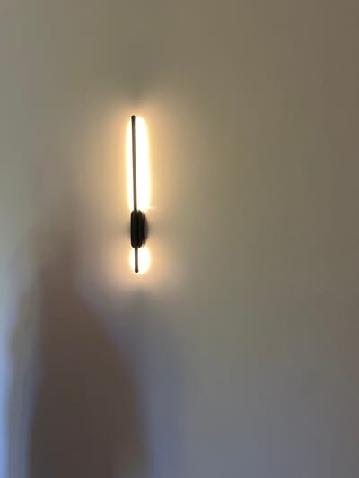A modern, black, vertical sconce light fixture with a thin, illuminated bar is mounted on a textured, light-colored wall. The light emitted is a warm yellow glow that casts a soft shadow to the left of the fixture. The fixture itself appears to be a single, thin vertical rod attached to a small, dark rectangular base with rounded edges, positioned in the center of the rod. The scene is static and devoid of any human presence. The environment suggests an indoor setting, likely a wall within a residential or commercial space.