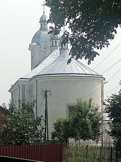A light-colored church with multiple domes, one large and two smaller, is visible from a slightly low angle, partially obscured by dark green foliage. The domes and the roof of the main building are covered in a metallic, silver material. A cross is visible atop the tallest dome. In the foreground, a brown fence runs horizontally across the bottom of the image. Behind the fence, there are trees with dense green leaves. A utility pole with wires is situated to the left of the church. The sky in the background is a hazy white. The church is the Church of the Holy Trinity in Yampil, Ukraine.
