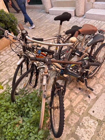 The image shows a collection of bicycles in Grad Rovinj, Croatia. Several bicycles are stacked or leaning against each other on a cobblestone surface. A person wearing blue jeans and white sneakers is partially visible on the left side of the image. Some greenery is visible on the right side of the frame.