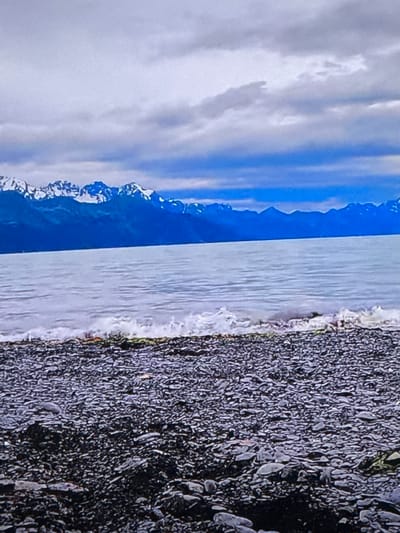 This is a medium shot, outdoor, landscape photograph. The scene depicts a body of water lapping against a rocky shore, with snow-capped mountains in the background under a cloudy sky. The image is composed of a foreground of dark, rounded rocks, a middle ground of a pale, rippling body of water, and a background of dark, imposing mountains topped with snow. The sky above is a mix of light and dark gray clouds, with some patches of blue peeking through. The overall tone of the image is cool and subdued, suggesting a cool or overcast day. No people or man-made objects are visible in the frame. The location is not identifiable as Springboro, United States of America.
