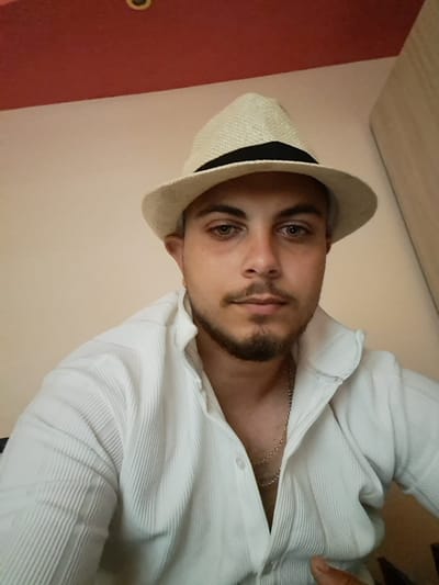The image is a selfie of a man wearing a straw hat with a black band, a white collared shirt, and a silver chain. He has light-colored eyes, dark hair, and a beard. The background is a wall painted in two tones, with a red section at the top and a beige section below. There is also a portion of a wooden closet visible on the right side of the frame. The photo appears to have been taken indoors, in a residential setting. Based on the provided information, the location is identified as Ignatievo, Bulgaria.