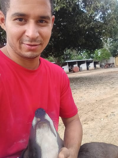 A man in a red t-shirt holds a dog with a black and white face. He is smiling at the camera. They are outside, with a dirt ground visible. There are some bushes and a structure in the background, possibly a building.