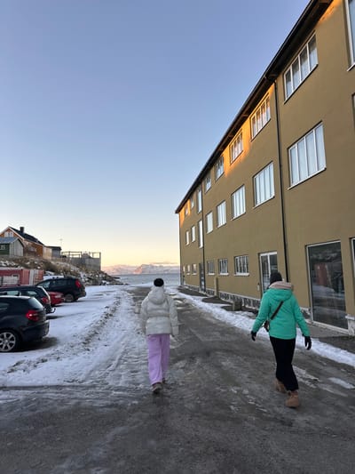 This image depicts a winter scene in Henningsvær, Norway, under a clear, pale blue sky. Two individuals walk away from the viewer on a partially snow-covered, gravel road that slopes gently towards a body of water and distant snow-capped mountains. The person on the left wears a white puffer jacket, light purple trousers, and a black beanie. The person on the right wears a teal puffer jacket, dark trousers, dark gloves, light brown boots, and carries a dark cross-body bag.
To the right, a three-story building with a muted olive-green or beige facade stands parallel to the road. It features multiple rectangular windows with white frames, some on the upper stories reflecting warm, golden sunlight. To the left, a snow-covered embankment hosts a small parking area with several vehicles, including a dark hatchback, a red sedan, and a dark SUV. Further left and slightly uphill, several traditional-style houses, one painted orange-brown, are visible. The background reveals a wide body of water, likely a fjord or bay, extending to the horizon where rugged, snow-dusted mountains rise under the soft, low-angle sunlight.