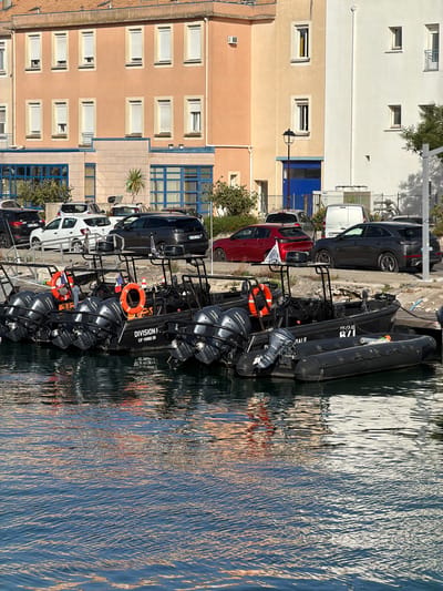 The image depicts a waterfront scene in Martigues, France, with a row of inflatable boats docked alongside a quay. The boats are equipped with multiple outboard motors and have orange life preservers. The backdrop consists of buildings with light-colored facades and terracotta roofs, typical of Mediterranean architecture. Several cars are parked on the quay behind the boats. The water is calm, with reflections of the boats and buildings visible on its surface. The overall impression is one of a functional port area, possibly used by a maritime service or organization.