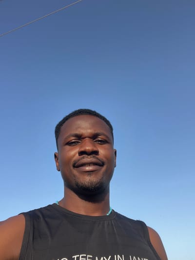 The media file is a portrait of a man against a clear blue sky, likely taken outdoors in Dam Camp, Nigeria. The man, who appears to be of African descent, is looking towards the camera with a slight smile. He is wearing a black sleeveless shirt with white text on it. The text, partially visible, reads "WHO TEE MY IN JANE". He also has a light blue beaded bracelet on his left wrist. The time of day appears to be daytime, with bright sunlight illuminating his face, suggesting fair weather. The low angle of the shot, looking up at the man against the sky, creates a sense of expansiveness and emphasizes the clear sky. There are no other people or objects prominently featured in the scene.